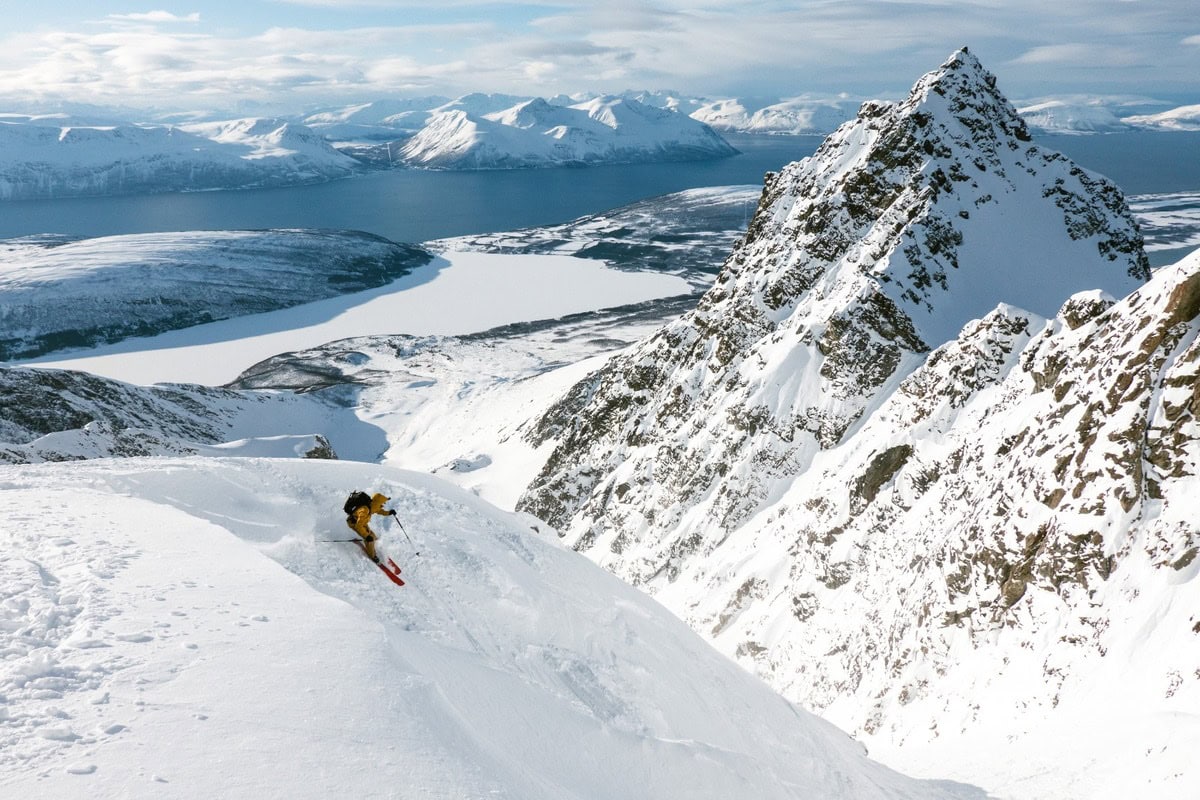 Skifahrer in gelber Jacke fährt einen Steilhang in den Lyngenalpen über dem Fjord ab