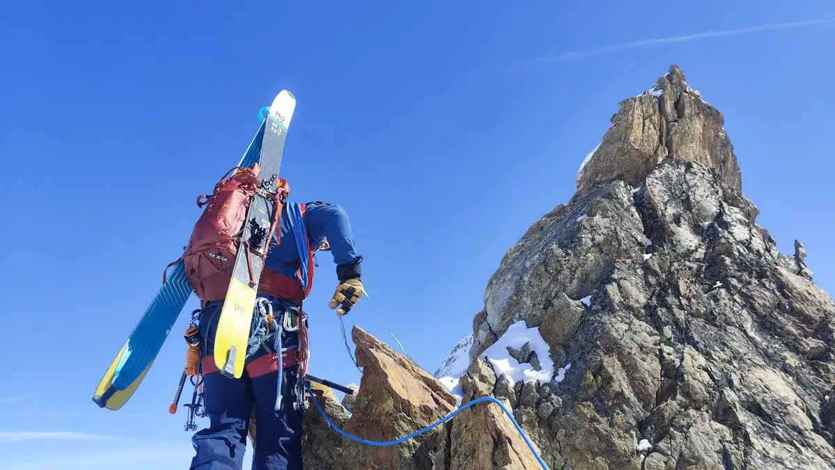 Bergsteiger mit Skiern am Rucksack auf einem Felsgrat vor einem Gipfelturm