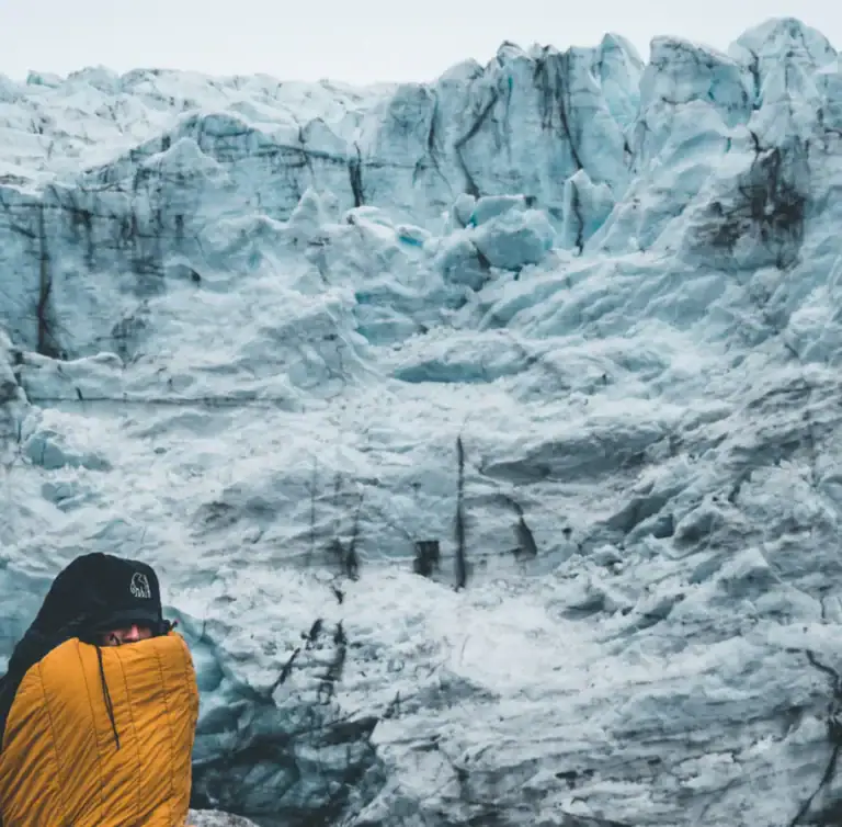 Person in gelber Jacke vor der zerklüfteten blauen Eiswand eines Gletschers